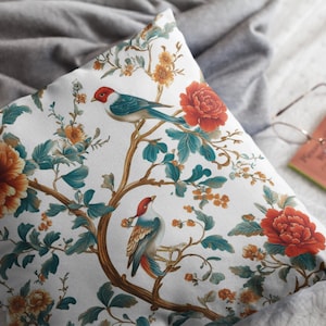 Puede incluir: Una almohada blanca con un patrón floral que presenta hojas verde azuladas, flores naranjas y rojas, y dos pájaros azules con acentos rojos.