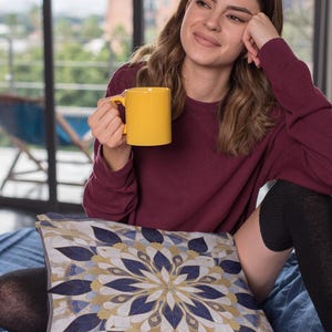 Puede incluir: Una mujer se sienta en una cama con una taza amarilla en la mano. Lleva un suéter marrón y leggings negros. La cama tiene una almohada de tiro con estampado azul y blanco.