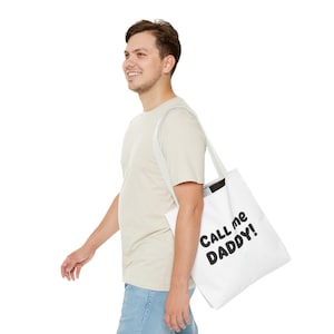 Puede incluir: Bolso tote blanco con el texto "CALL me DADDY!" en letras negras en negrita. El bolso se lleva al hombro. La persona de la imagen lleva una camiseta de color claro y vaqueros azules.