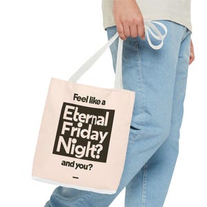 Puede incluir: Un bolso tote de color rosa claro con asas blancas. El bolso presenta el texto "Feel like a Eternal Friday Night? and you?" en negro y negrita. El bolso lo sostiene una persona que lleva vaqueros azul claro.