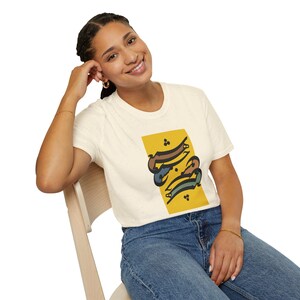 Puede incluir: Una camiseta de manga corta color crema con un gráfico rectangular amarillo en el pecho. El gráfico contiene letras estilizadas de estilo caligráfico en marrón oscuro, azul y verde con pequeños puntos negros dispuestos verticalmente.