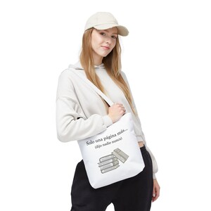 Puede incluir: Bolso tote blanco con el texto "Solo una página más... (dijo nadie nunca)" y un gráfico de libros apilados. El bolso tiene una correa para el hombro y lo lleva una persona que lleva una sudadera con capucha y una gorra color crema.
