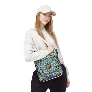 Puede incluir: Un bolso tote con un vibrante e intrincado diseño floral en tonos azules, dorados y turquesas. El bolso tiene una correa blanca y se lleva al hombro. El fondo es blanco.