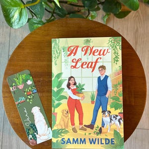 May include: A green and white bookmark with a dog and plants on it, next to a book titled "A New Leaf" by Samm Wilde. The book cover features a couple with dogs in a plant shop.