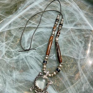 Puede incluir: Un collar con un colgante de tortuga plateada y una piedra transparente. El collar tiene un cordón negro y está adornado con cuentas de madera y plata. El colgante de tortuga mide unos 3,8 cm de largo.