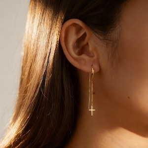 May include: Gold-colored dangle earrings featuring a small cross pendant. The earrings have a delicate chain design that hangs from a hoop. The cross pendant is a simple, elegant shape.