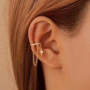 Op de afbeelding: Gouden oor manchet met een rij kleine heldere stenen en bungelende kettingen. Een stervormige bedel met een centrale heldere steen hangt aan de manchet. De sieraden worden op het oor gedragen.