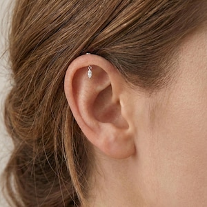 May include: A close-up of an ear with a small, dangling earring. The earring features a teardrop-shaped, clear gemstone suspended from a delicate chain. The earring is silver-toned and adds a subtle sparkle. The ear is set against a backdrop of light brown hair.