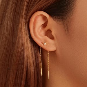Puede incluir: Primer plano de un conjunto de pendientes dorados. El perno superior presenta una pequeña gema cuadrada y transparente. Delicadas cadenas doradas cuelgan, cada una terminando en una fina barra dorada.