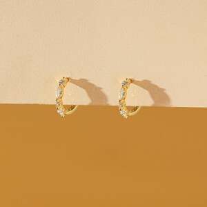 May include: A pair of gold-colored hoop earrings. Each earring is adorned with a series of clear, sparkling gemstones. The earrings are set against a warm, neutral background, highlighting their design and color.