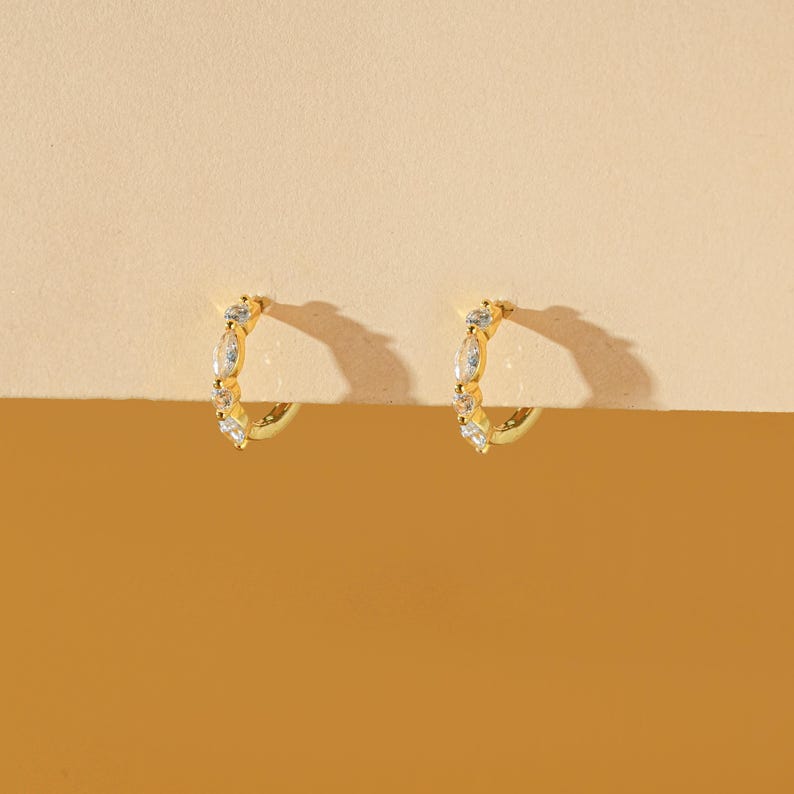 May include: A pair of gold-colored hoop earrings. Each earring is adorned with a series of clear, sparkling gemstones. The earrings are set against a warm, neutral background, highlighting their design and color.