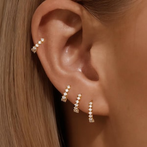 May include: Gold-toned ear jewelry set featuring a curved ear cuff and three hoop earrings, each adorned with small, sparkling gemstones. The jewelry is displayed on an ear, showcasing the design and fit.