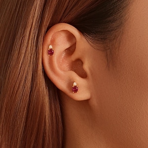 Puede incluir: Primer plano de una oreja adornada con dos pendientes de oro. Cada pendiente presenta una piedra preciosa roja oscura en forma de lágrima, acentuada con un pequeño diamante brillante. Los pendientes están engarzados en oro.