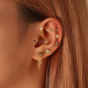 Puede incluir: Una oreja adornada con múltiples pendientes dorados con varios diseños. Los pendientes incluyen pequeños tachuelas, pendientes de cadena colgantes y piezas con piedras preciosas verdes y formas de hojas. La oreja está colocada sobre un fondo de cabello castaño claro.