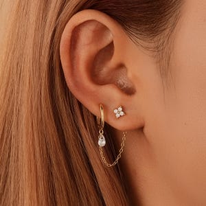 Puede incluir: Primer plano de una oreja adornada con pendientes dorados. Se ve un pequeño pendiente con forma de flor, junto con un pendiente de aro con un dije en forma de lágrima. Una delicada cadena conecta el pendiente de aro con el pendiente.