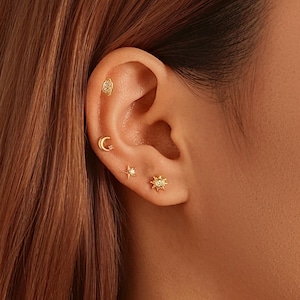 May include: An ear featuring several gold earrings. The collection includes a small oval stud, a crescent moon, a star with a central gem, and a sunburst design. The earrings are displayed against the skin of the ear and surrounding area, showcasing the jewellery.
