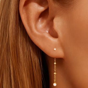 May include: A close-up of a person's ear adorned with two gold earrings. One is a small stud, and the other is a dangling earring with a chain of stars and a small, round, white charm at the end. The earrings are set against the backdrop of the person's skin and hair.