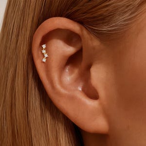 Puede incluir: Un piercing dorado con tres piedras preciosas claras de corte cuadrado. La joya está colocada en una línea vertical en el cartílago de la oreja, con la oreja y el pelo en el fondo.