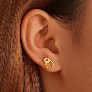 Puede incluir: Pendientes de botón dorados con un diseño de doble círculo. Un círculo es liso, mientras que el otro está adornado con pequeñas piedras brillantes. Los pendientes se muestran en una oreja, destacando su tamaño y estilo.