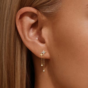May include: Gold-coloured star and drop earrings. The earrings have a star-shaped stud with small clear stones and a dangling chain with two clear, round stones. The earrings are worn on a person's ear.