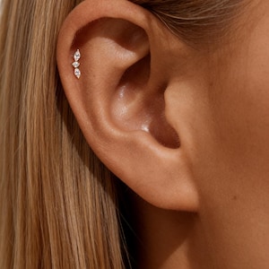 Può includere: Un piercing all'orecchio in oro con tre gemme chiare a forma di goccia. Il gioiello è posto verticalmente sull'orecchio, con le gemme di dimensioni graduate. L'orecchio è visibile con capelli biondi sullo sfondo.