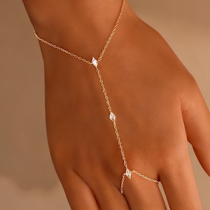 Peut inclure: Une fine chaîne de main dorée ornée de trois pierres précieuses claires en forme de diamant. La chaîne est reliée à une bague et à un bracelet, créant un accessoire élégant. Le bijou est présenté sur une main, sur fond neutre.