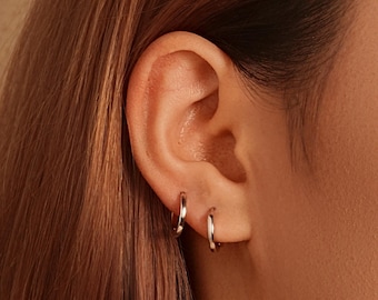 May include: Close-up of an ear with two small, silver hoop earrings. The earrings are simple and shiny, reflecting light. The skin tone is light, and the hair is a warm brown color.
