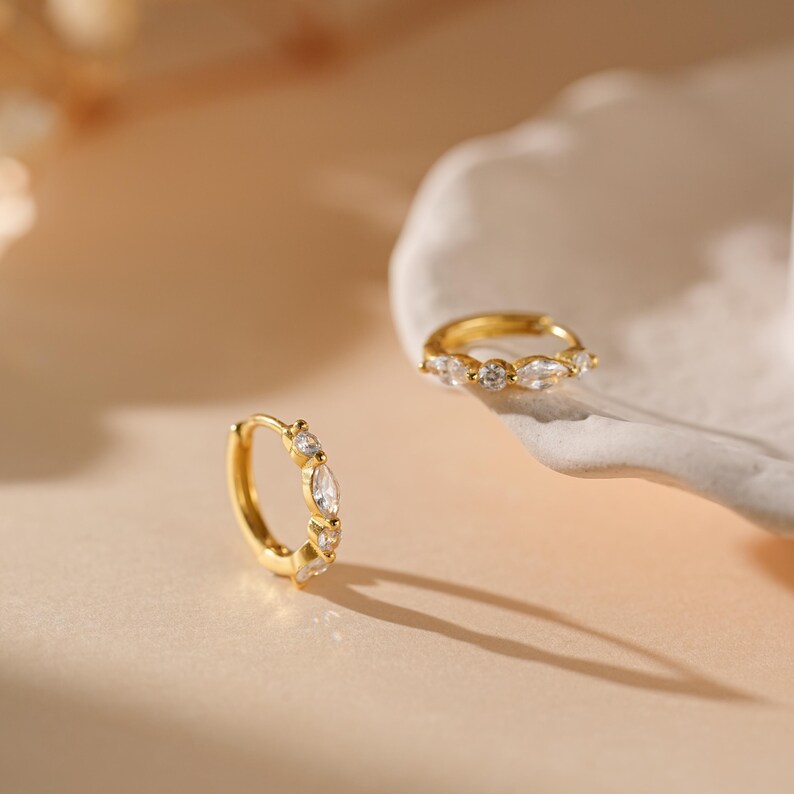 May include: Two gold-colored hoop earrings with clear, marquise-cut and round-cut gemstones. One earring is in focus, while the other is slightly out of focus. The earrings are displayed on a light-colored surface with a white, shell-shaped dish.
