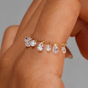Puede incluir: Un delicado anillo de oro con una cadena con gemas claras en forma de lágrima colgantes. El anillo se lleva en un dedo, mostrando el intrincado diseño y las piedras brillantes. La joya es elegante y adecuada para varias ocasiones.