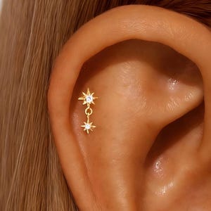 May include: A gold-colored ear piercing featuring two star-shaped charms, one dangling below the other. Each star has a central, clear gemstone. The earring is set against the backdrop of a person's ear and skin.