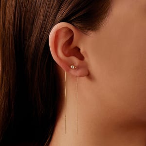May include: A gold earring featuring a small square gemstone and a fine chain that hangs down. The earring is worn on an ear. The person has brown hair.