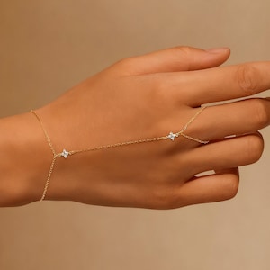 Puede incluir: Una delicada cadena de mano dorada con detalles en forma de flor de cuatro pétalos, cada uno con pequeñas piedras preciosas transparentes. La cadena se conecta a un anillo y se envuelve alrededor de la mano, creando una joya elegante y minimalista.