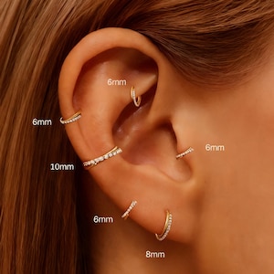 Puede incluir: Primer plano de una oreja adornada con múltiples pendientes dorados. Los pendientes incluyen pequeños aros y barras, algunos con piedras transparentes incrustadas. Tamaños: 6mm, 8mm y 10mm. La oreja tiene un tono de piel claro.