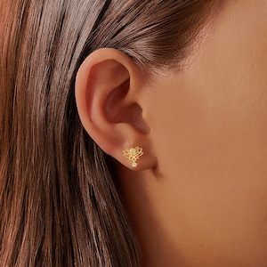 Könnte beinhalten: Nahaufnahme eines goldfarbenen Lotusblüten-Ohrrings mit einem kleinen, herabhängenden Akzent. Der Ohrring wird am Ohr einer Person getragen. Der Ohrring ist ein zartes Schmuckstück.