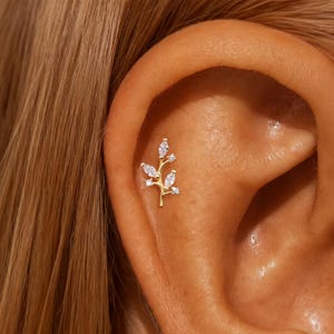 Puede incluir: Un pendiente dorado con un delicado diseño de rama. La rama está adornada con varias piedras preciosas transparentes con corte marquesa, creando una estética brillante inspirada en la naturaleza. El pendiente está colocado en la oreja de una persona.