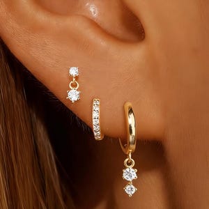 Może przedstawiać: Zestaw trzech kolczyków w kolorze złotym. Jedna para to małe koło z wiszącym przezroczystym kamieniem szlachetnym. Kolejny to mały sztyft z wiszącym kamieniem szlachetnym. Trzeci to małe koło z przezroczystymi kamieniami szlachetnymi.