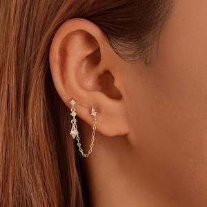 May include: Gold-toned ear jewelry featuring a chain connecting two earrings. One earring has a small stud with a diamond, and the other has a dangling design with diamonds. The earrings are worn on a person's ear.