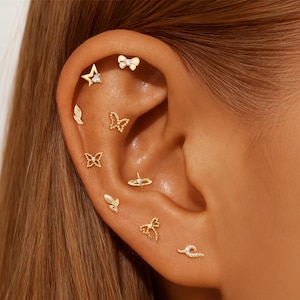 May include: A close-up of an ear adorned with multiple gold-colored earrings. The earrings feature various designs, including a star, bow, butterfly, leaf, and other delicate shapes. Some earrings have small, clear gemstones. The ear is set against a background of light brown hair.