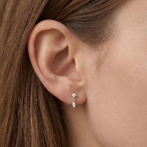 May include: A close-up of a person's ear with a small, dangling earring. The earring features a round, clear gemstone at the top, connected to a teardrop-shaped gemstone below. The earring is silver-colored and set against the skin.