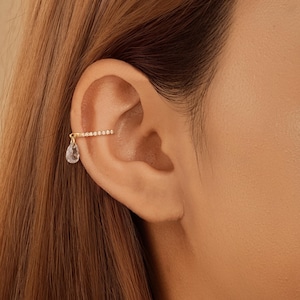 May include: Gold-toned ear cuff featuring a row of small, clear crystals along the top and a dangling teardrop-shaped crystal. The jewelry is worn on the ear, showcasing its delicate design and sparkle.