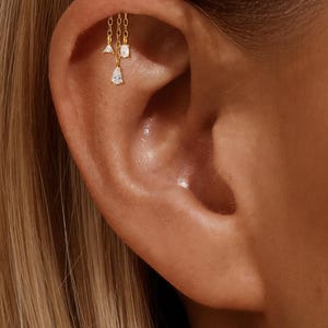 Op de afbeelding: Een goudkleurige oorbel met een hangend kettingontwerp. De ketting bevat drie kleine, heldere, edelsteenachtige bedels: een driehoek, een rechthoek en een druppelvorm. De sieraden worden op het oor gedragen.