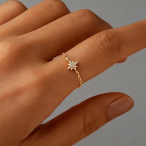 May include: A delicate gold chain ring with a starburst design. The starburst is embellished with small, sparkling gemstones. The ring is worn on a finger, highlighting its elegant and minimalist style. The ring's design is suitable for everyday wear.