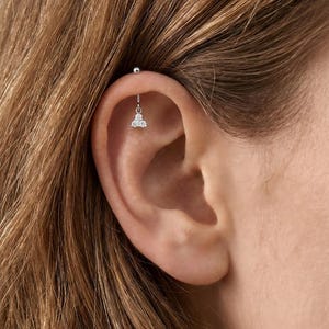 May include: A close-up of an ear with a silver industrial barbell earring. The barbell has a small ball on one end and a dangling charm with three small, clear gemstones. The ear is set against a backdrop of light brown hair.