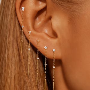 May include: Close-up of multiple gold earrings with dangling chains and clear gemstones. The earrings feature various designs, including teardrops, stars, and geometric shapes. The jewelry is worn on a person's ear, showcasing the delicate and elegant style.