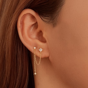 May include: Gold-toned double earrings with a star and square-cut cubic zirconia design. The earrings are connected by a delicate chain, with a small star charm dangling below. The earrings are shown on an ear.