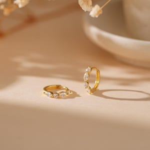 May include: Two gold-colored hoop earrings with clear, marquise-cut stones. The earrings are displayed on a light-colored surface, with a soft, neutral background. The earrings are a jewelry item.