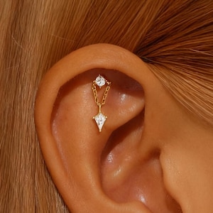 May include: A gold-colored ear piercing with a dangling design. The piercing features a small, round, clear gemstone at the top, connected by a delicate chain to a larger, diamond-shaped gemstone. The jewelry is set against the backdrop of a person's ear.