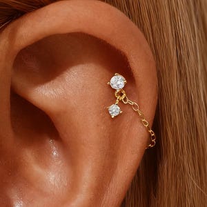 Könnte beinhalten: Ein goldfarbener Ohrschmuck mit zwei klaren, rund geschliffenen Edelsteinen. Der obere Edelstein ist am Ohr befestigt, und eine zarte Kette verbindet ihn mit einem zweiten, kleineren Edelstein darunter. Der Schmuck befindet sich vor dem Hintergrund eines Ohrs.