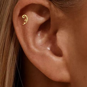 Pode incluir: Um close-up de uma orelha com um pequeno brinco dourado. O brinco tem um design curvo e decorativo. A orelha está colocada sobre um fundo de cabelo castanho claro.