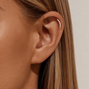 May include: A curved ear cuff featuring a row of small, sparkling clear gemstones. The jewelry is set against the backdrop of a person's ear and hair, with a soft, neutral background.